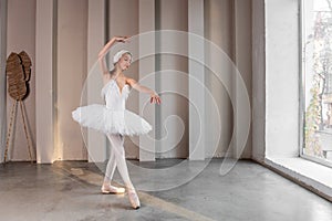 Elegant young ballerina performs beside large windows, poised stance, delicate white tutu