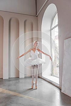 Elegant young ballerina performs beside large windows, poised stance, delicate white tutu
