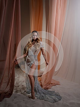 An elegant young blonde in a luxurious dress behind a curtain