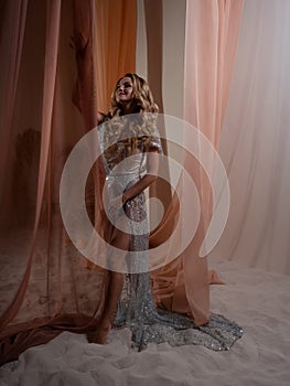 An elegant young blonde in a shiny translucent dress behind a curtain