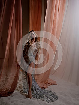 An elegant young blonde in a shiny translucent dress behind a curtain