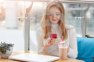 Elegant young blonde woman in white sweater sits on blue sofa in cafe, holds pink mobile phone and surfes internet or types mesage