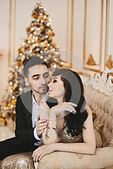 An elegant young couple in luxurious outfits sits next to a Christmas tree. New Year party