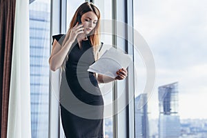 Elegant young female business owner holding documents talking on mobile phone discussing contract conditions standing in