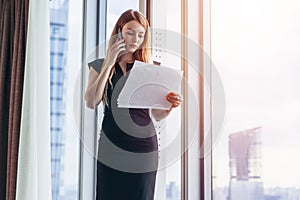 Elegant young female business owner holding documents talking on mobile phone discussing contract conditions standing in