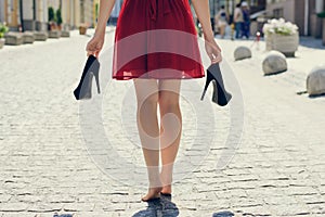 Elegant young girl in red dress with high-heels in hands, walking on the street barefoot. She is coming back home after party in photo