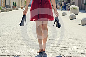 Elegant young girl in red dress with high-heels in hands, walking on the street barefoot. She is coming back home after party in