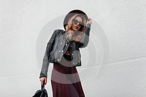 Elegant young hipster woman in a luxury hat in sunglasses in a leather jacket in pants with a backpack with a cape traveling