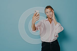 Elegant young lady in casual wear trying to take selfie while holding smartphone, isolated on blue
