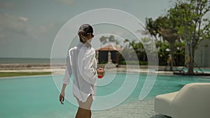 Elegant young lady enjoying good day near the hotel pool, wearing white shirt holding glass of red juice with straw