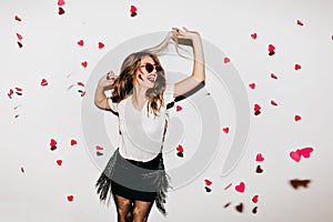 Elegant young lady in trendy sunglasses looking up at fallen hearts. Portrait of positive long-haired woman in short