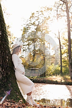 Elegant young pregnant woman in white knit dress and hat touching and stroking belly, relaxing in park by the lake on