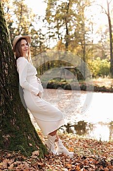Elegant young pregnant woman in white knit dress and hat touching and stroking belly, relaxing in park by the lake on