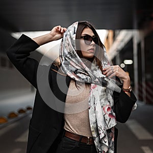 Elegant young pretty beautiful woman in stylish dark sunglasses in a black coat puts a luxurious silk scarf on head outdoors.