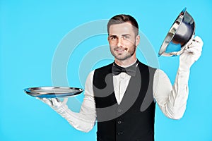 Elegant young waiter holding cloche over empty tray ready to serve on blue background