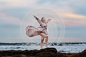 Elegant young woman in beautiful long dress waving in the wind with a deep neckline stay on ocean beach on the rock on sunset