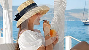 Elegant young woman in big hat and long dress drinking cocktail with straw while relaxing in gazebo on summer sea resort