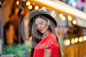 Elegant young woman in a red dress and a black hat on a background of street cafe lights