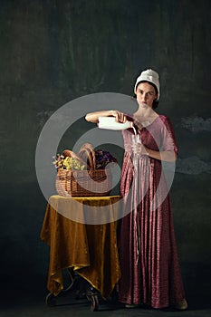 Elegant young woman in renaissance dress, maid pouring milk into glass against dark vintage background. Peasant girl