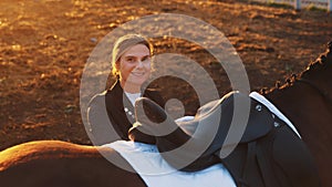 an elegant young woman with a smile on her face stands next to a brown horse on uneven ground