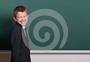 Elementary school boy make faces near blank chalkboard background, dressed in classic black suit, one pupil, education concept