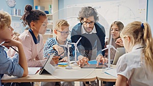 Elementary School Classroom: Enthusiastic Teacher Holding Tablet Computer Explains Lesson to Brill