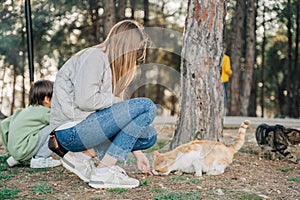 Elementary school kid and his mother petting feeding a stray cat in the city park. Mom and son child boy playing with