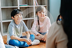 Elementary school kids sitting on the floor and looking at teacher in their classroom. The teacher is reading story tale from a