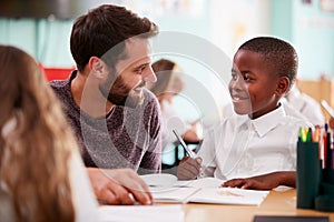 Elementary School Teacher Giving Male Pupil Wearing Uniform One To One Support In Classroom