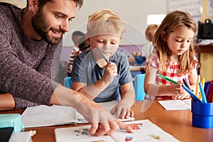 Elementary School Teacher Helping Pupils As They Work At Desk In Classroom