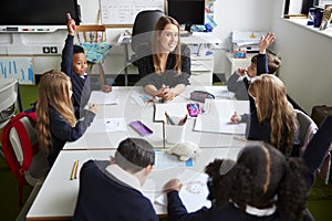 Elevated view of female primary school teacher sitting at a table in a classroom with schoolchildren raising their hands during a