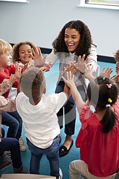 Elevated view of infant school children in a circle in the classroom giving high fives to their smiling female teacher, vertical,