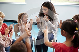 Elevated view of infant school children sitting on chairs in a circle in the classroom, raising hands and learning to count with t