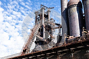 Elevated walkway alongside steel mill blast furnaces against a blue sky with clouds