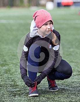 Eleven years old girl football player makes tying shoelaces on cleats, artificial green field