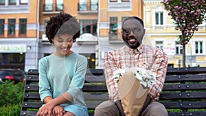 Embarrassed couple sitting on bench with flowers, first date, affection emotions