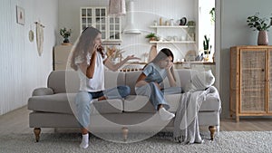Embittered mother is talking on phone and pointing finger to side of sad daughter sits on couch