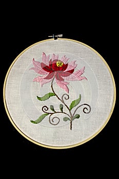 Embroidered flower with red and pink petals on twisted branches and leaves on white cotton fabric with a wooden frame on a black b