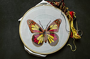 Embroidered  bright butterfly on white cotton fabric stretched on a wooden Hoop with colored threads and scissors lying on black p