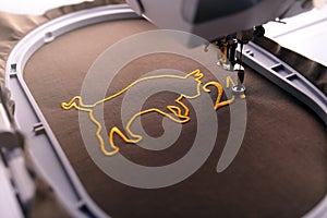 Embroidery hoop and needle foot of an embroidery machine stitching ox symbol and number 21 with golden yarn on shiny fabric