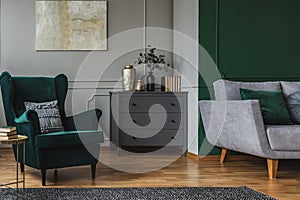 Emerald green armchair with pillow next to grey wooden commode in dark living room interior