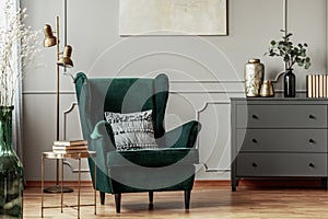 Emerald green armchair with pillow next to grey wooden commode in dark living room interior