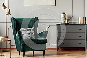Emerald green wing back chair in grey living room interior with wooden commode