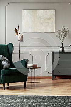 Emerald green wing back chair with pillow in grey living room interior with wooden commode