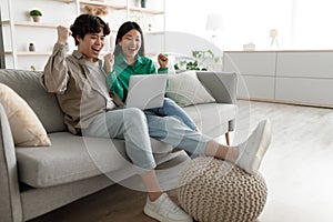 Emotional Asian couple with laptop making YES gesture, celebrating success, sitting on couch at home, free space