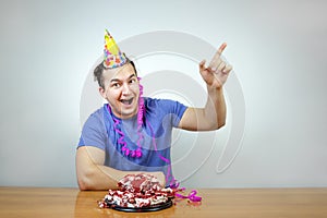 Emotional beautiful caucasian man with birthday party cone hat on head and crumple cake raises your index finger up, smiling happi