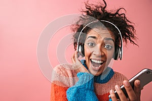 Emotional beautiful young african woman posing isolated over pink wall background listening music with headphones