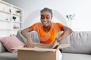 Emotional black female customer receiving long-awaited package, opening carton box at home, satisfied with purchase