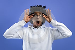 Emotional black teenager opening mouth in shock, touching eyeglasses, looking at camera on blue studio background