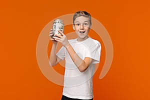 Emotional boy holding glass money box on orange background